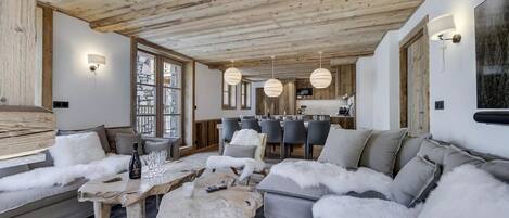 A cozy living room with a wooden ceiling, a stone-framed window, and a dining area with a wooden table and chairs.