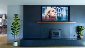 A modern living room with a large flat-screen TV mounted on a dark wall, a fireplace below, and two potted plants on either side. - Stylish Mid-Century Modern Home - Near DT Portland for your perfect getaway! (Milwaukie)