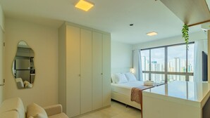 Une chambre d’hôtel moderne comprenant un lit, un coin salon avec un canapé, une grande fenêtre donnant sur la ville et un miroir au mur.