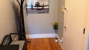 Un pasillo con piso de madera, una silla a cuadros blanco y negro, una silla de metal negro, un cuadro colgado en la pared con un avión, un jarrón con flores amarillas y una puerta blanca.