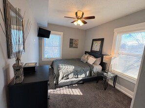 A bedroom with a bed, a ceiling fan, a TV, and two windows with blinds.