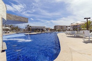 A swimming pool with a curved edge, surrounded by lounge chairs and umbrellas, and a building in the background. - Muro Alto Club - Ground Floor Apto by Carpediem (Ipojuca)