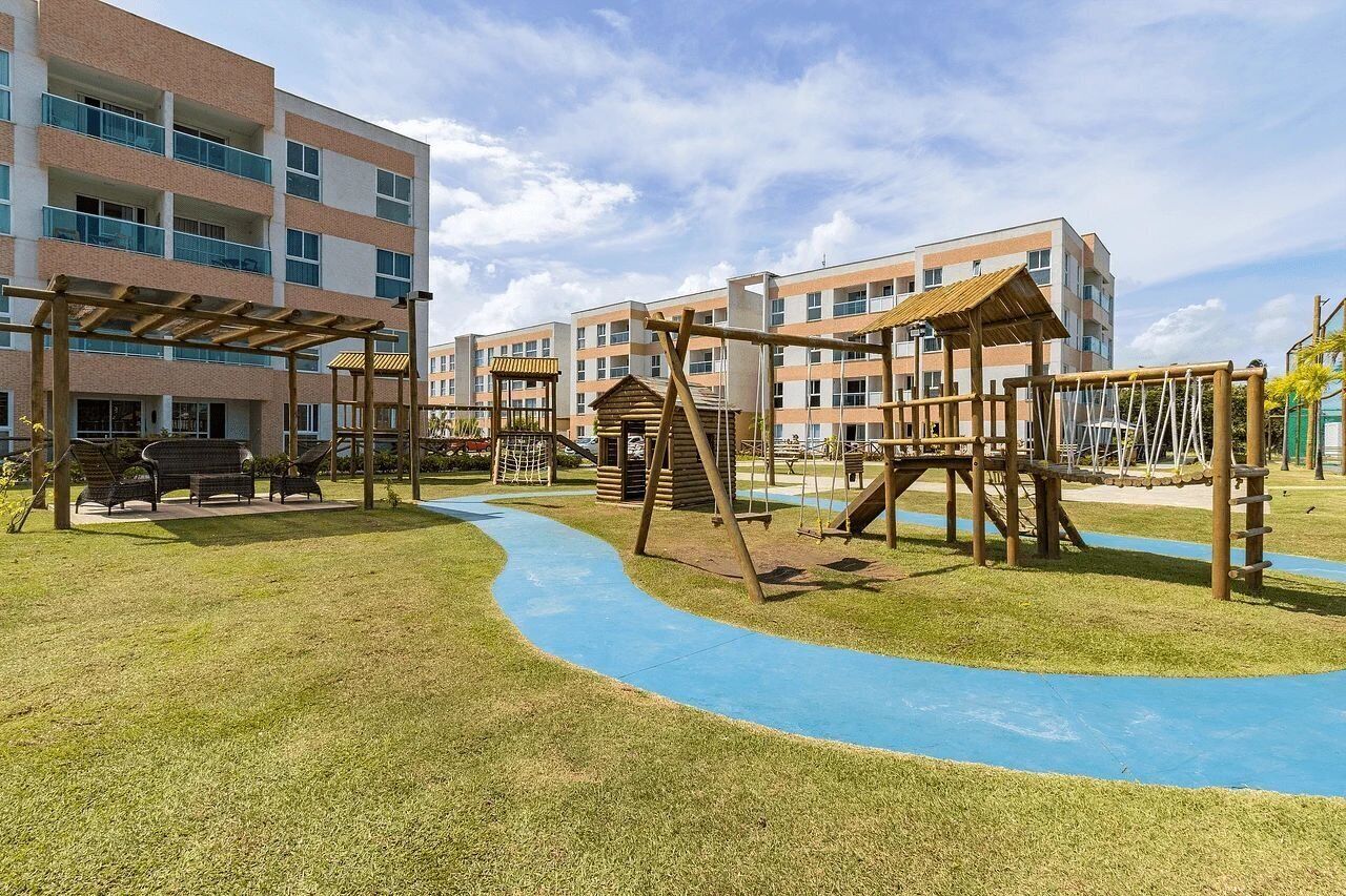 A playground with a blue rubberized surface, wooden play structures, and a building with multiple balconies in the background.