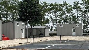 Una hilera de edificios modulares grises con ventanas pequeñas, un estacionamiento pavimentado y un primer plano con césped.
