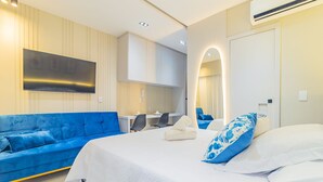 A modern hotel room with a large bed, a blue sofa, a desk, and a flat-screen TV. - Makia Beach #c22 - Apto Inteligente by Carpediem (Ipojuca)