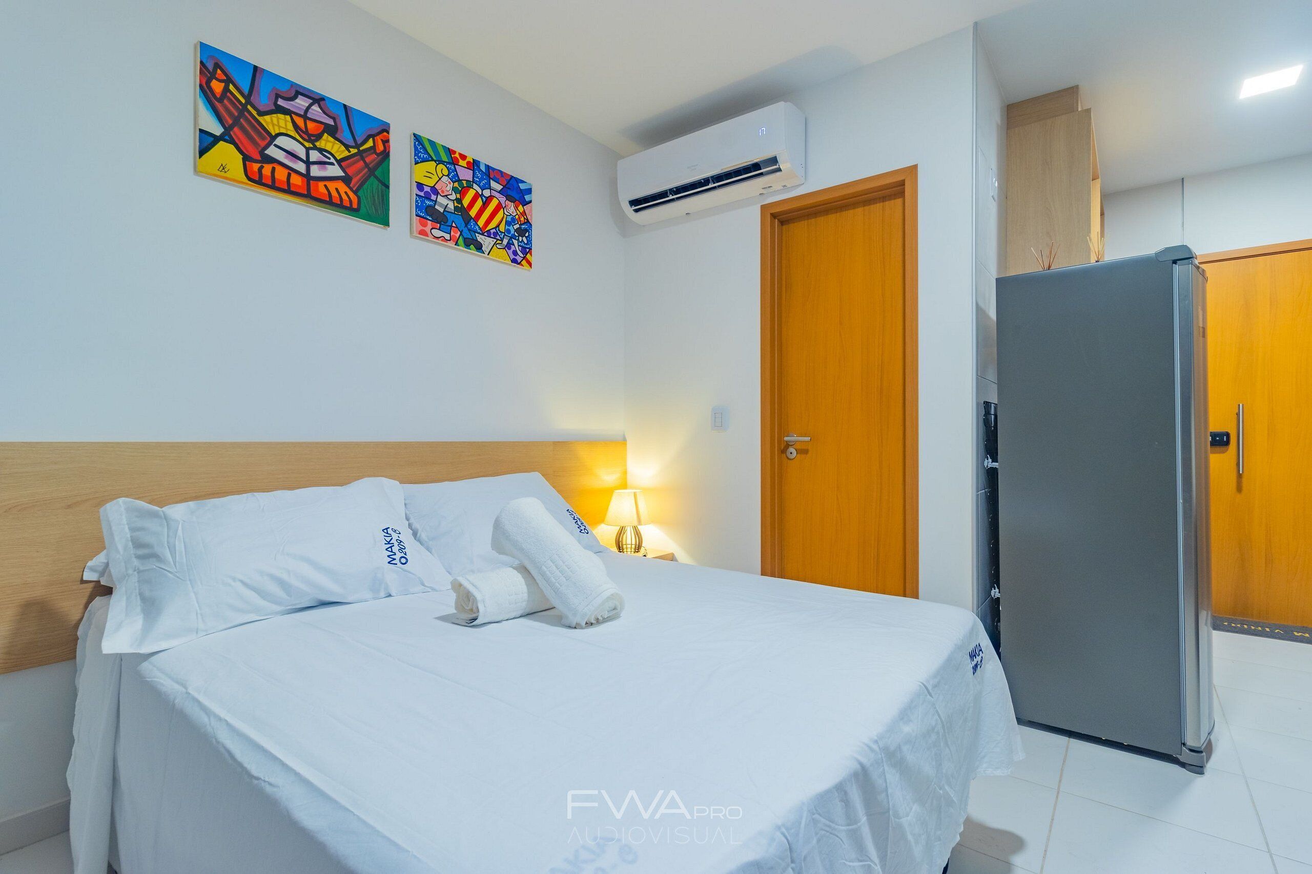 Habitación de hotel con cama, ropa de cama blanca, cabecera de madera, aire acondicionado empotrado, refrigerador y dos cuadros coloridos.