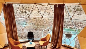 Une pièce aménagée comme une tente, avec un plancher en bois, deux fauteuils orange, une petite table sur laquelle sont posées des bouteilles, et une vue sur un paysage rocheux grâce à de grandes fenêtres.
