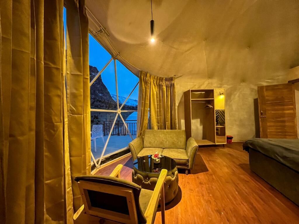 A cozy room with a sofa, a small table, and a chair. There is a window with curtains, a bed, and a wardrobe.