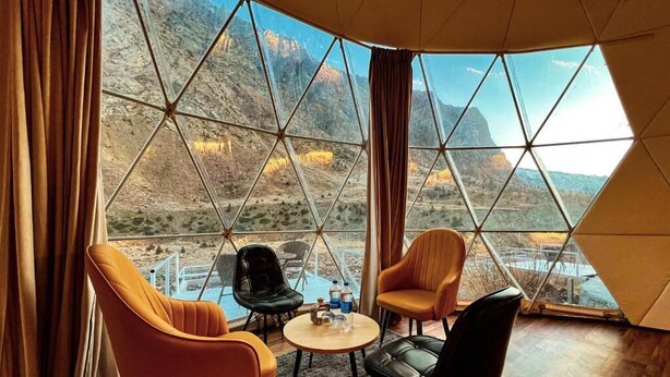 Une pièce dotée d’une grande fenêtre donnant sur un paysage montagneux. À l’intérieur, on trouve deux fauteuils orange et un fauteuil noir disposés autour d’une petite table ronde sur laquelle se trouvent une bouteille et des verres.