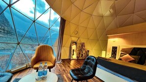 Un intérieur moderne avec une grande fenêtre en forme de dôme de verre, un canapé, deux fauteuils, une petite table ronde avec des bouteilles et un plancher en bois.