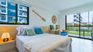 A modern hotel room with a large bed, a nightstand, a lamp, a view of the building outside, and a decorative wall hanging. - Makia Beach #D3 - em Muro Alto por Carpediem (Ipojuca)