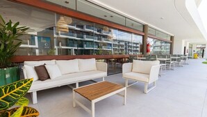 A modern outdoor seating area with white sofas and tables, a glass railing, and a view of a building with balconies. - Makia Beach #D3 - em Muro Alto por Carpediem (Ipojuca)