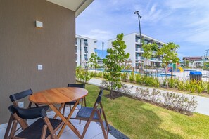 Een terras met een ronde houten tafel en stoelen, een kleine boom, en uitzicht op een zwembad en gebouwen op de achtergrond.