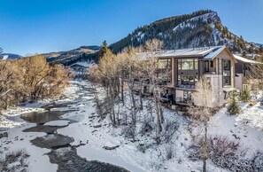 A modern lodge with a glass facade, situated by a snow-covered river, with mountains in the background.