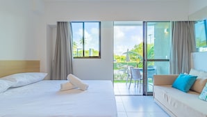 Quarto de hotel com cama, sofá, varanda com mesa e cadeiras, e vista para áreas verdes e edifícios.