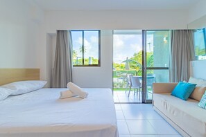 Ein Hotelzimmer mit Bett, einer Sitzecke, einem Balkon mit Tisch und Stühlen sowie Blick auf Grünflächen und Gebäude.