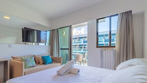 A hotel room with a bed, a sofa, a TV, and a balcony with a view of buildings. - Makia Beach #d205 - Flat in Ipojuca by Carpediem (Ipojuca)
