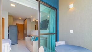 A modern hotel room with a glass door leading to a balcony, a dining area with a table and chairs, a sofa, and a refrigerator. - Makia Beach #d205 - Flat in Ipojuca by Carpediem (Ipojuca)