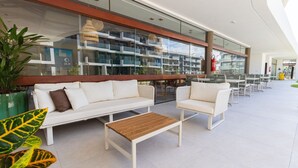 A modern outdoor seating area with white sofas and tables, a glass railing, and potted plants. - Makia Beach #d205 - Flat in Ipojuca by Carpediem (Ipojuca)