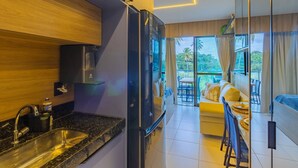 Eine moderne Küche mit integriertem Kühlschrank, Spüle und Kaffeemaschine. Es gibt einen Essbereich mit Tisch und Stühlen, sowie ein Sofa. Der Raum verfügt über große Fenster mit Blick auf Palmen.