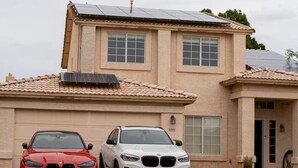 A two-story house with a garage, two cars parked in front, and a solar panel on the roof.