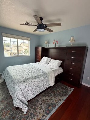 Un dormitorio con cama, ventilador de techo, ventana y cómoda.