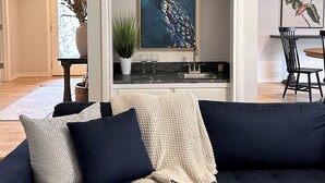 A modern living room with a dark blue sofa, a framed peacock painting, a potted plant, and a dining area with a table and chairs. - Spacious Hilltop Retreat in Upscale Cul-de-Sac Neighborhood (Tulsa)