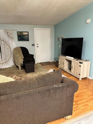 Ein Wohnzimmer mit einem braunen Sofa, einem Fernseher auf einer Holzkommode, einer Tür und einem Fenster mit Seeblick.