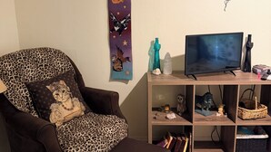 Een woonkamer met een fauteuil in luipaardprint, een televisie op een houten standaard, en een wand met een decoratief wandkleed.