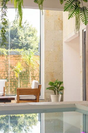 A modern indoor pool area with a clear view of the outdoors, including a brick wall and greenery. There are two wooden chairs and a potted plant near the pool. - Briones House (xalapa)