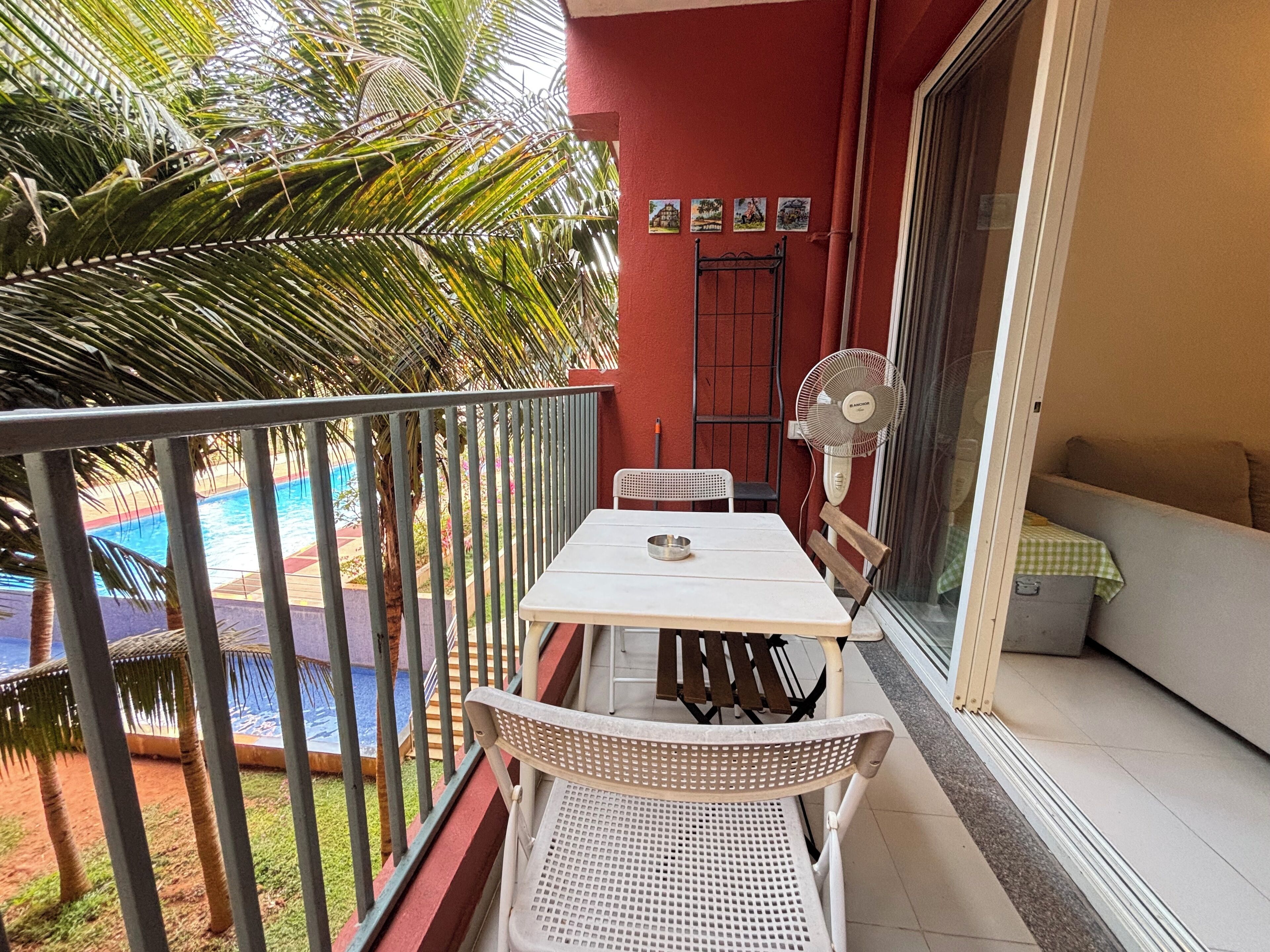 A balcony with a table, chairs, a fan, and a view of a swimming pool.