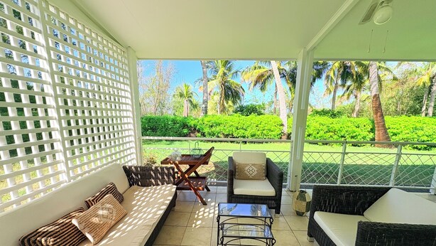 A covered outdoor seating area with wicker furniture, a glass table, and a view of a well-maintained garden with palm trees. - Seaside villa in the middle of a coconut grove Saint Leu (Saint-Leu)