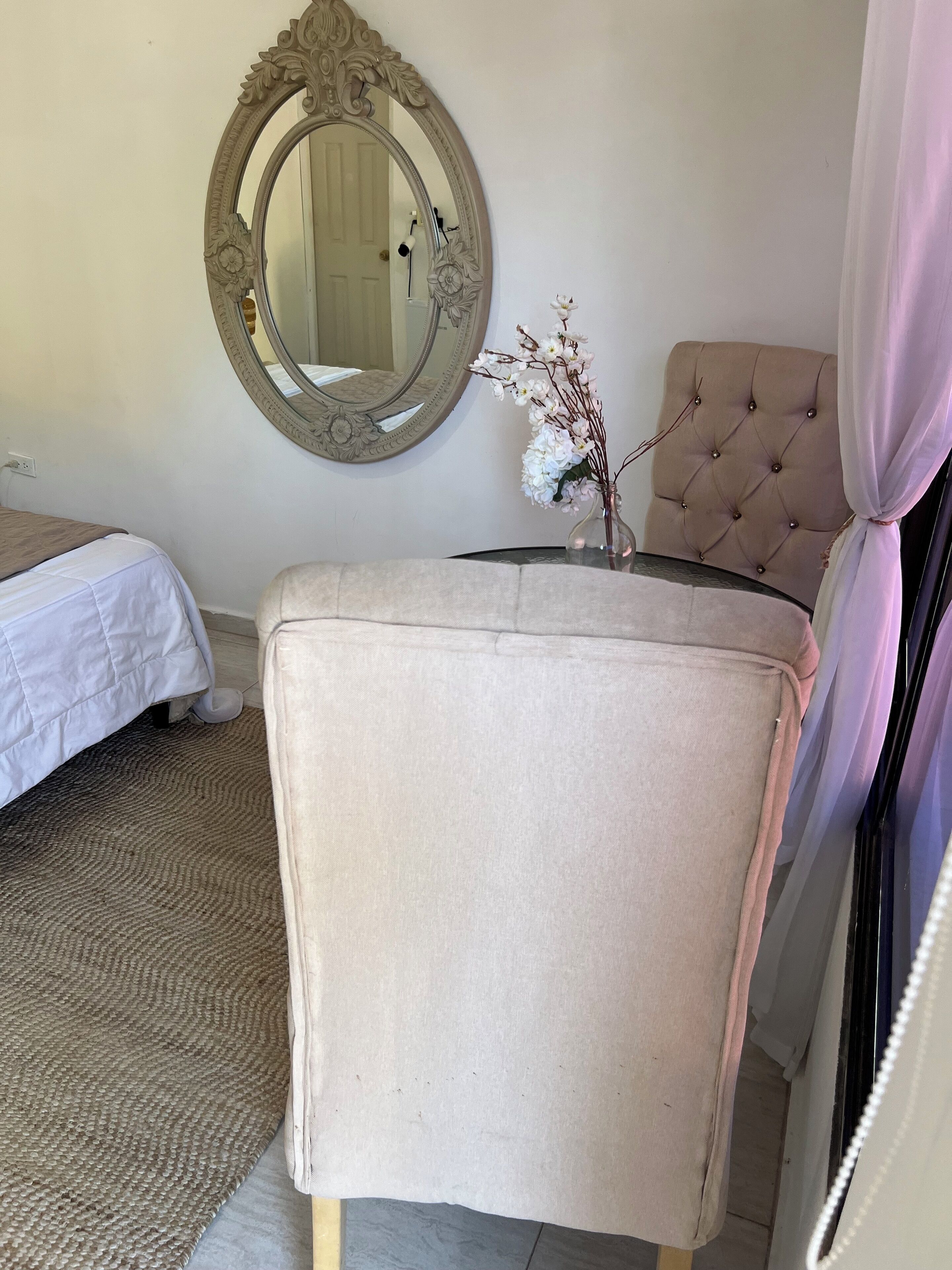Un dormitorio con cama, cabecera capitonada, espejo y un florero con flores.
