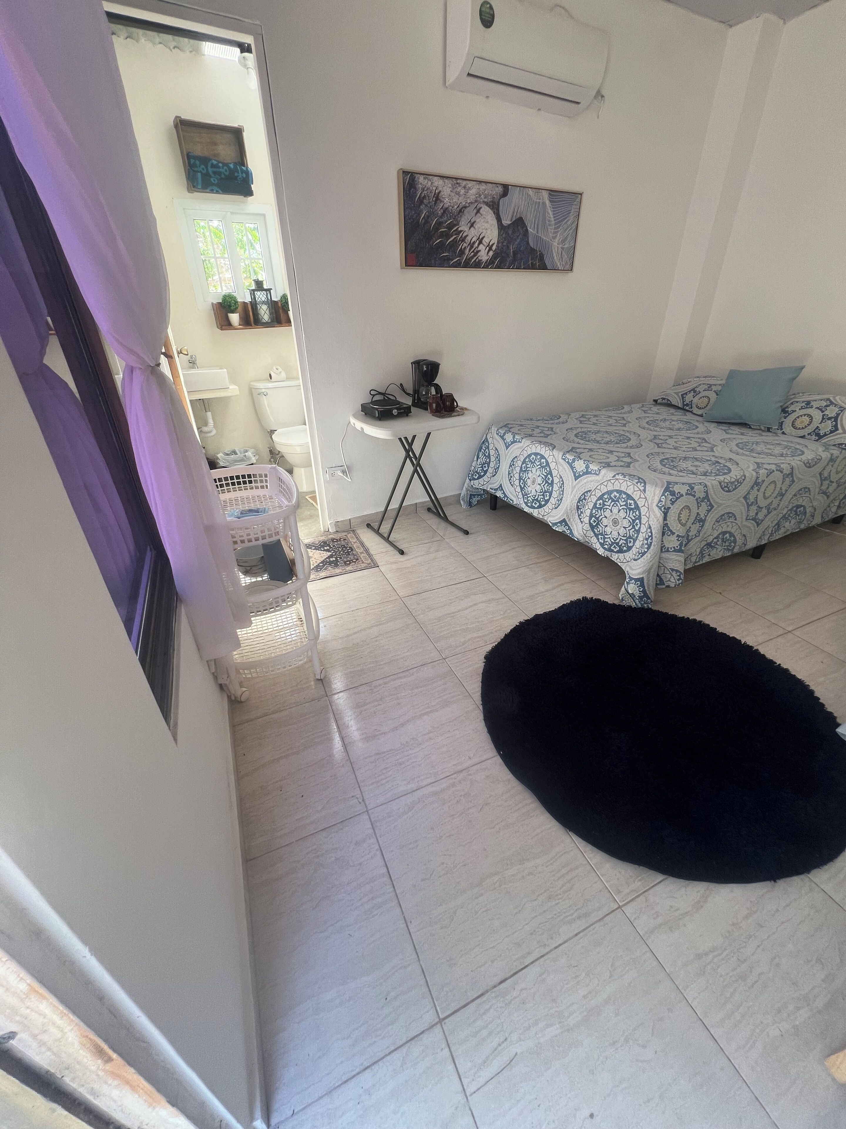 A bedroom with a bed, a small table with a coffee maker, a black furry rug, and a painting on the wall.