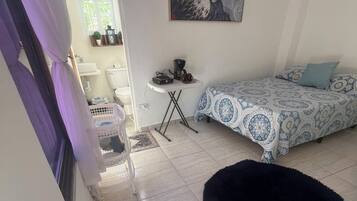 A bedroom with a bed, a small table with a coffee maker, a black furry rug, and a painting on the wall.