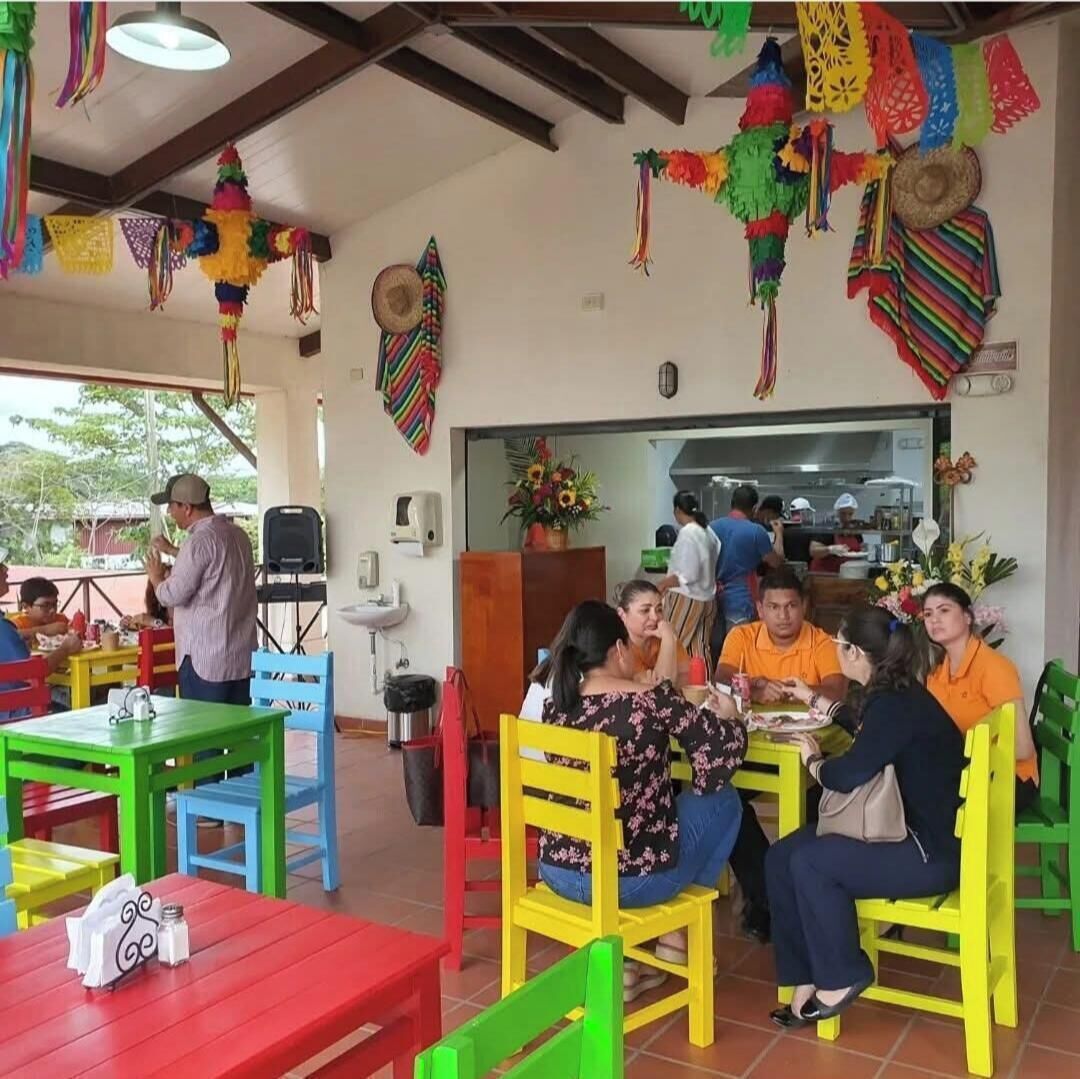 Ein Restaurant mit bunten Stühlen und Tischen, ein Mann macht ein Foto und im Hintergrund ist eine Küche zu sehen.