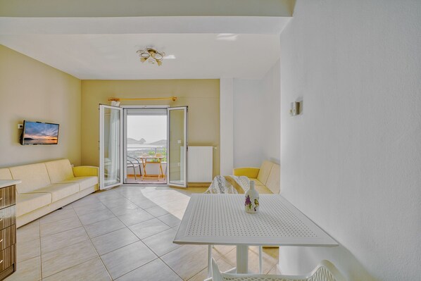 A bright, modern living room with a sofa, a dining table, and a TV on the wall. - NIKOLA APARTMENTS (Lemnos)