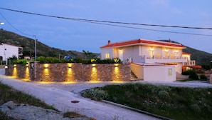 A two-story house with a red roof, a stone wall with lights, and a driveway. - NIKOLA APARTMENTS (Lemnos)