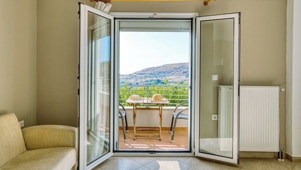 A room with a sliding glass door leading to a balcony with a table, chairs, and a view of a hilly landscape. - NIKOLA APARTMENTS (Lemnos)