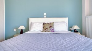 A neatly made bed with a quilted white cover, two bedside lamps, a floral pillow, and a wall socket. - NIKOLA APARTMENTS (Lemnos)