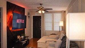 Ein Wohnzimmer mit Fernseher, der 'NETFLIX' anzeigt, Deckenventilator, Sofa, Lampe und einer Tür mit der Aufschrift 'THE ALANO'.