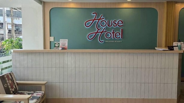 Un mostrador de recepción con un letrero que dice 'House Hotel' y 'Walking Street'. A la izquierda, una pequeña mesa con una silla y un cuadro enmarcado.