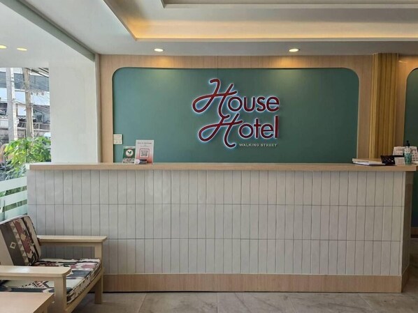 Un mostrador de recepción con un letrero que dice 'House Hotel' y 'Walking Street'. A la izquierda, una pequeña mesa con una silla y un cuadro enmarcado.