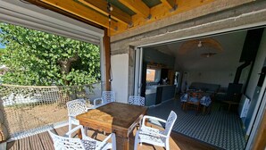 En overdækket terrasse med et træbord og hvide stole, et køkkenområde kan anes gennem en åben dør, og der er udsigt til grøn vegetation udenfor.