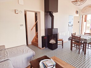A living room with a sofa, a dining table with chairs, a black wood-burning stove, and a staircase leading to another room. - Holiday Home “La Maison De L'écume” with Terrace, Garden & Wi-Fi (La Tranche-sur-Mer)
