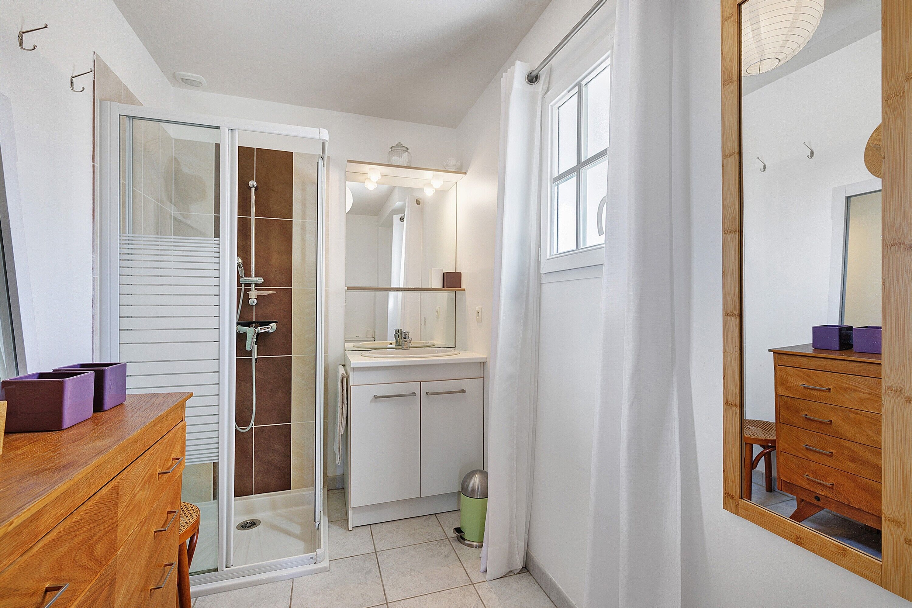 Een badkamer met een douchecabine van glas, een houten wastafelmeubel, een spiegel en een klein afvalbakje.