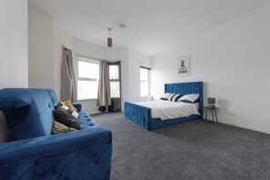 A bedroom with a blue bed, a blue armchair, and a small table with a lamp.
