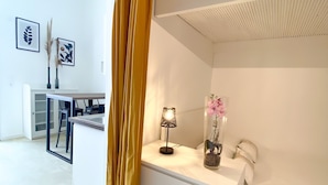 Ein modernes Schlafzimmer mit einem weißen Bett, einem Nachttisch mit Lampe, einer Vase mit Blumen und einem Schreibtisch mit Stuhl.
