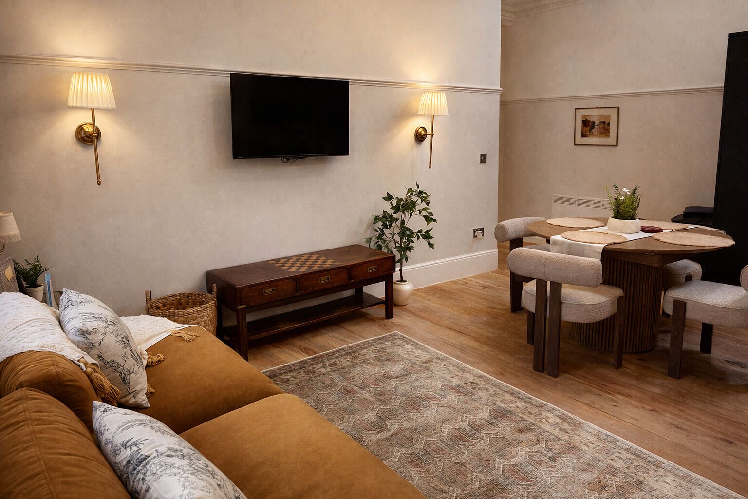A modern living room with a sofa, a wooden coffee table, a dining table with chairs, a TV mounted on the wall, and a plant.
