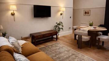 A modern living room with a sofa, a wooden coffee table, a dining table with chairs, a TV mounted on the wall, and a plant.